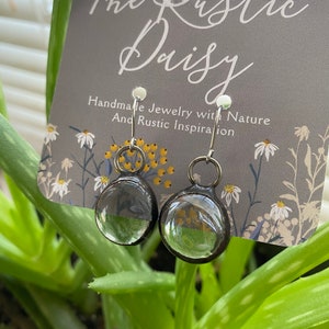 May include: A pair of clear glass earrings with silver hoops and black metal accents. The earrings are displayed on a card with the text "The Rustic Daisy Handmade Jewelry with Nature and Rustic Inspiration".