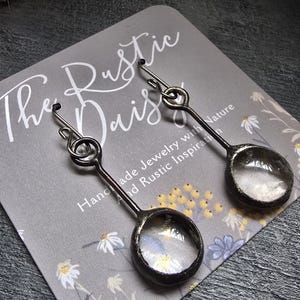 May include: A pair of silver earrings with a clear glass circle pendant. The earrings are handmade and have a rustic, nature-inspired design. The card reads "The Rustic Daisy Handmade Jewelry with a Rustic Inspiration".