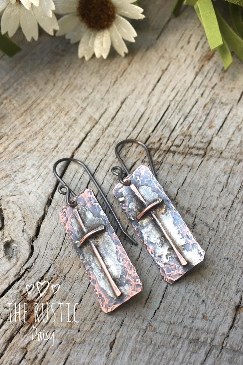 Puede incluir: Un par de pendientes de cobre martillado con un dise&ntilde;o de cruz. Los pendientes son de color plateado y cobre y tienen un aspecto r&uacute;stico.