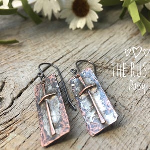 Puede incluir: Un par de pendientes de plata y cobre con textura martillada y dise&ntilde;o de cruz. Los pendientes cuelgan de una superficie de madera. El texto "THE RUSTIC Daisy" es visible en el fondo.