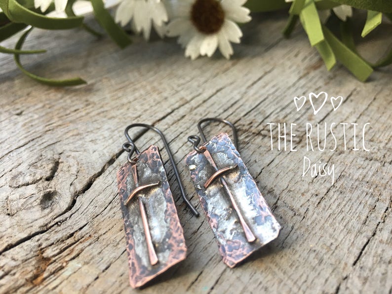 Puede incluir: Un par de pendientes de plata y cobre con textura martillada y dise&ntilde;o de cruz. Los pendientes cuelgan de una superficie de madera. El texto "THE RUSTIC Daisy" es visible en el fondo.