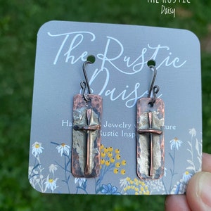 Puede incluir: Un par de pendientes con un dise&ntilde;o de cruz de cobre martillado y plata. Los pendientes est&aacute;n en una tarjeta que dice "The Rustic Daisy" y "Joyer&iacute;a con inspiraci&oacute;n r&uacute;stica".
