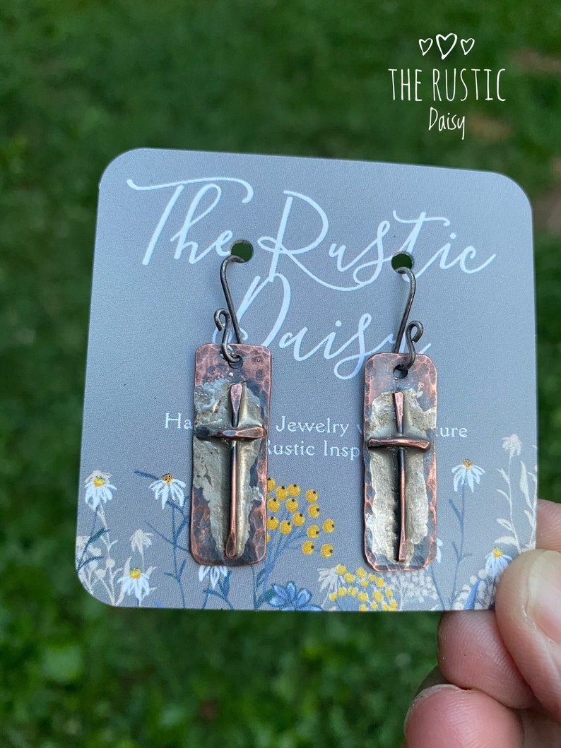 Puede incluir: Un par de pendientes con un dise&ntilde;o de cruz de cobre martillado y plata. Los pendientes est&aacute;n en una tarjeta que dice "The Rustic Daisy" y "Joyer&iacute;a con inspiraci&oacute;n r&uacute;stica".