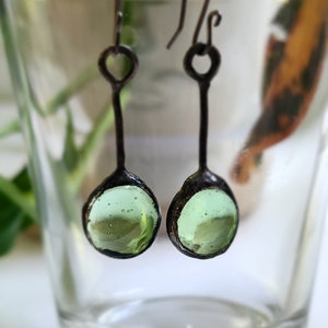 May include: A pair of green glass and silver earrings with a teardrop shape. The earrings have a simple design with a small loop at the top and a round green glass pendant.