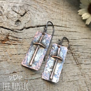 Puede incluir: Un par de pendientes de plata y cobre con textura martillada y dise&ntilde;o de cruz. Los pendientes cuelgan de una superficie de madera.