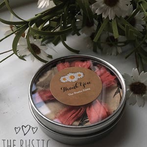Puede incluir: Una peque&ntilde;a lata de plata con una tapa transparente que contiene una cinta rosa y un c&iacute;rculo de papel marr&oacute;n con el texto "Thank You The Rustic Daisy".