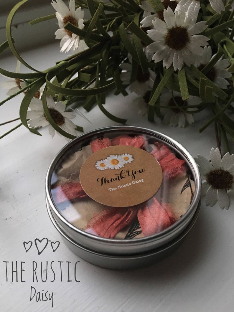 Puede incluir: Una peque&ntilde;a lata de plata con una tapa transparente que contiene una cinta rosa y un c&iacute;rculo de papel marr&oacute;n con el texto "Thank You The Rustic Daisy".