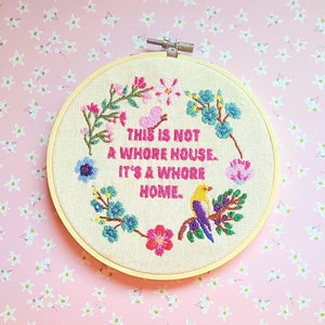 May include: A circular wooden embroidery hoop containing beige fabric with hand-stitched floral motifs, a small bird, and the pink embroidered text: THIS IS NOT A WHORE HOUSE. IT'S A WHORE HOME. The design features colorful flowers and greenery.