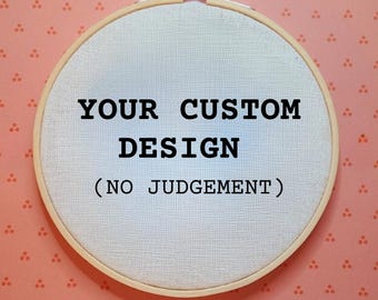 Könnte beinhalten: Ein runder Stickrahmen aus Holz, der einen cremeweißen Stoff einspannt. Auf dem Stoff befindet sich der schwarze Schriftzug "YOUR CUSTOM DESIGN (NO JUDGEMENT)" in einer Schreibmaschinen-Schriftart. Der Rahmen hat oben eine Metallschraube.