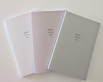 May include: Three notebooks in shades of white, lavender, and gray are arranged side by side. Each notebook has the words "pause. write. be." printed in a minimalist font. The notebooks are a simple, elegant design.