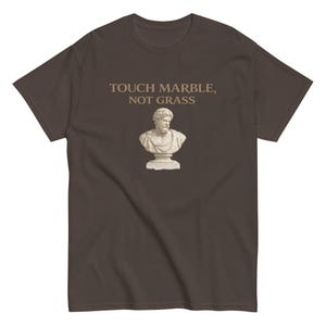 May include: A dark brown short-sleeve t-shirt featuring a graphic of a classical white marble bust of a bearded man. Above the bust, the text reads "TOUCH MARBLE, NOT GRASS" in a light tan, serif font centered on the chest of the garment.