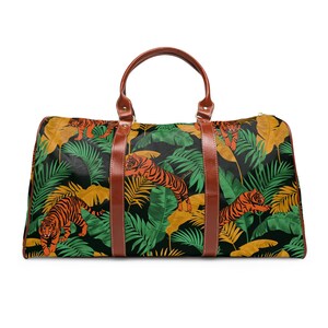 Peut inclure: Un grand sac de voyage avec un imprimé jungle, représentant des tigres oranges, des feuilles vertes et jaunes, sur fond noir. Il possède des poignées et des détails en cuir marron, et une fermeture éclair dorée.
