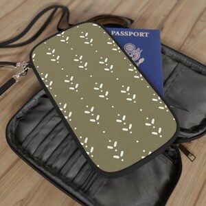 May include: Olive green travel wallet with a white leaf pattern and black trim. The wallet is open, revealing a blue passport. A black strap and zipper are visible.