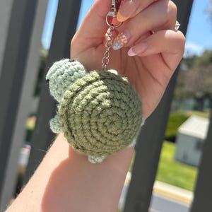 May include: A handmade crochet turtle keychain in shades of green. The turtle has a textured shell and small, light green head and feet. It is attached to a silver-colored keychain. The keychain is held up against a blurred outdoor background.