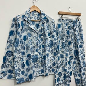 May include: A two-piece pajama set featuring a white background with a blue floral pattern. The set includes a button-down shirt with a chest pocket and matching drawstring pants. The pajamas are hanging on wooden hangers.