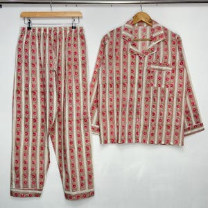 Pode incluir: Um conjunto de pijama de duas peças com um padrão de riscas florais. O conjunto inclui uma camisa de manga comprida com botões e bolso no peito e calças com cordão. O padrão apresenta riscas verticais com detalhes florais vermelhos sobre um fundo creme.
