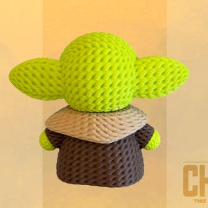 May include: A handmade green and brown crocheted figure of The Child, with large ears and a brown robe. The figure is made of textured yarn and has a tan collar. The text "THE CHILD" and "THIS IS THE WAY" is visible.