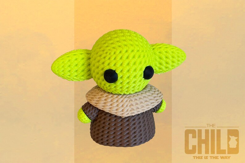 May include: A crocheted figure of The Child, featuring a lime green head with large ears and black eyes. It wears a tan and brown outfit. The text "THE CHILD THIS IS THE WAY" is in the lower right corner.