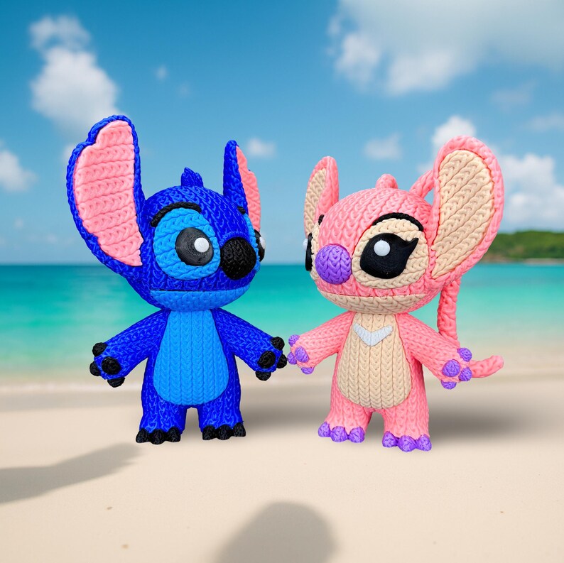 May include: Two knitted figurines of the characters Stitch and Angel from the animated film Lilo & Stitch. Stitch is blue with pink ears, and Angel is pink with beige accents. They are standing on a sandy beach with a blue ocean and sky in the background.