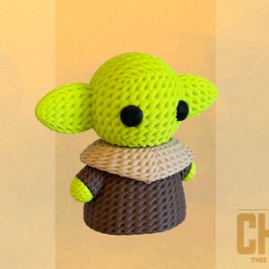 May include: A crocheted figure of The Child, featuring large green ears, black eyes, and a brown robe. The figure has a tan collar and is set against a light yellow background. The text "THE CHILD THIS IS THE WAY" is visible.