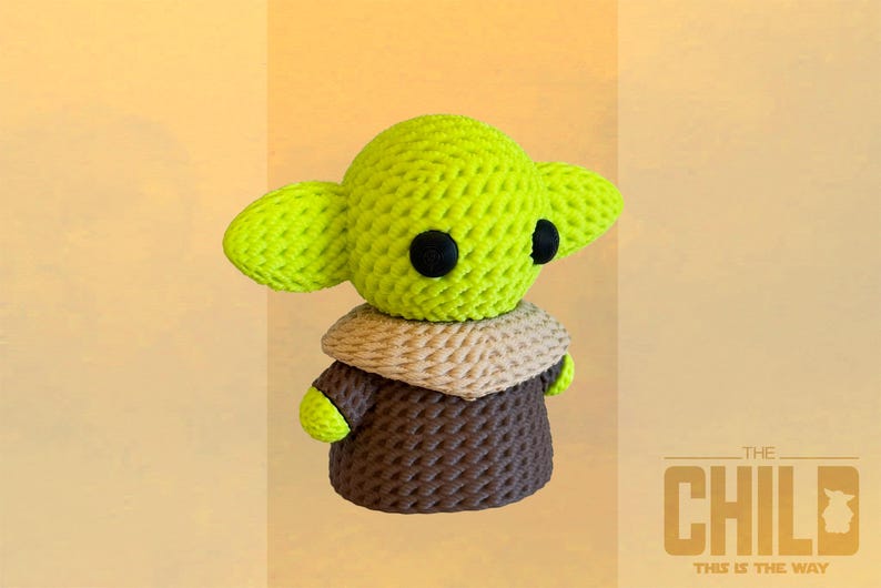 May include: A crocheted figure of The Child, featuring large green ears, black eyes, and a brown robe. The figure has a tan collar and is set against a light yellow background. The text "THE CHILD THIS IS THE WAY" is visible.
