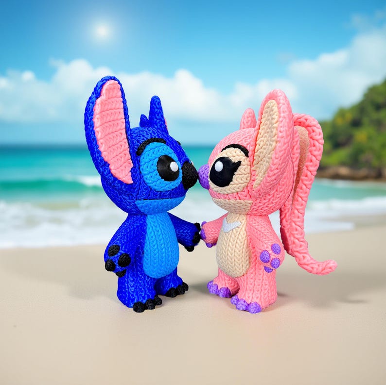May include: Two knitted character figurines, one blue and one pink, stand on a sandy beach with a blue sky and ocean in the background. The blue figurine has large ears and a friendly expression, while the pink figurine has long ears and a sweet face. They are holding hands.