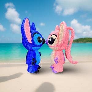 May include: Two knitted figurines of Stitch and Angel from Lilo & Stitch. Stitch is blue with pink accents, and Angel is pink with cream and purple details. They are positioned nose-to-nose against a backdrop of a beach and blue sky.