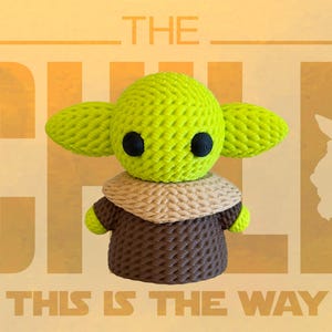 May include: A handmade green and brown crocheted figure of The Child, with large ears and black eyes. The figure is wearing a brown robe and a tan collar. The background is a light orange with the words "THE CHILD" and "THIS IS THE WAY".