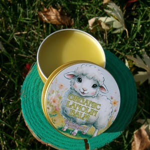 May include: An open, round, gold-colored tin of organic lanolin balm. The lid features a cartoon sheep with the text "Organic Lanolin Balm" and "Garden Harvest Apothecary". The balm is a light yellow color.