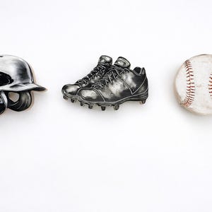 May include: A set of three pins featuring a black and white baseball batting helmet, a pair of black baseball cleats with detailed laces and studs, and a white baseball with prominent red stitching, all arranged in a horizontal row.