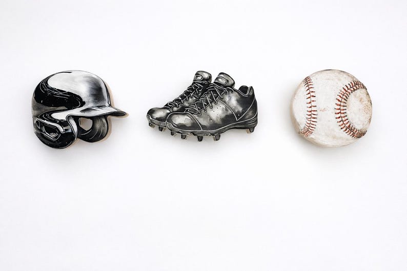 May include: A set of three pins featuring a black and white baseball batting helmet, a pair of black baseball cleats with detailed laces and studs, and a white baseball with prominent red stitching, all arranged in a horizontal row.