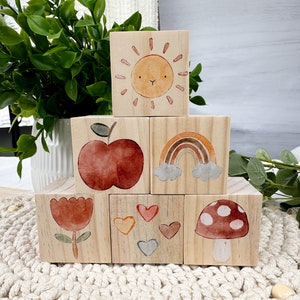 May include: Six wooden blocks with painted images of a sun, rainbow, apple, flower, hearts, and mushroom. The blocks are stacked in a pyramid shape.