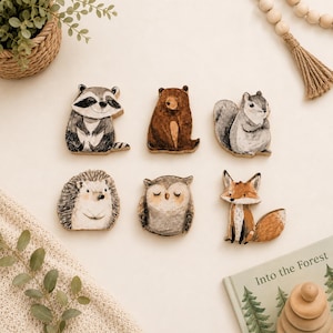 May include: A set of six wooden animal cutouts featuring a raccoon, a brown bear, a gray squirrel, a hedgehog, an owl with closed eyes, and a fox. Each figure is painted with detailed features and sits in a flat, decorative arrangement.
