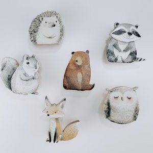 May include: A collection of six woodland animal figurines, including a hedgehog, raccoon, squirrel, bear, fox, and owl. Each figurine is painted in soft, natural colors and has a whimsical design. Perfect for nursery decor.