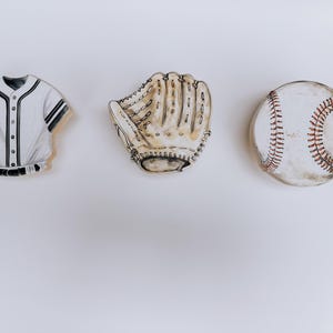 May include: A watercolour illustration of baseball equipment. It features a white and black baseball jersey, a tan baseball glove, and a white baseball with red stitching, set against a white backdrop.