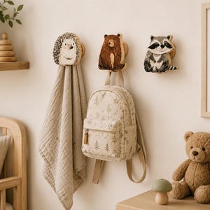May include: Three wooden wall hooks shaped like a hedgehog, a brown bear, and a raccoon. The hedgehog hook holds a beige muslin cloth, the bear hook supports a beige backpack with a forest print, and the raccoon hook is mounted empty.