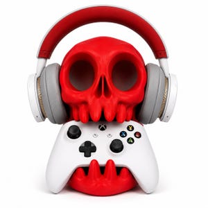 Puede incluir: Un soporte con forma de calavera de color rojo brillante que sostiene un mando de videojuego blanco. La calavera lleva unos auriculares de diadema blancos y rojos con almohadillas grises. El mando tiene una cruceta negra y dos joysticks.