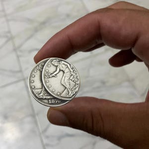 Puede incluir: Una moneda de plata con una mujer con túnica, una luna creciente y estrellas. La moneda está fechada en 1870. La moneda se sostiene entre dos dedos.