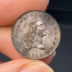 May include: A silver coin featuring a profile of a woman with flowing hair, the word "LIBERTY" above, and the date "1794" below. The coin's edge is lined with stars, and it appears to be an antique or collectible item.