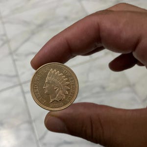 May include: A gold-colored 1859 Indian Head penny. The coin features a profile of a Native American in a feathered headdress on one side and the words "UNITED STATES OF AMERICA" on the other. The coin is held between two fingers.