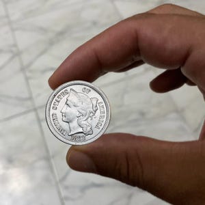 May include: A silver coin from the United States of America, held between fingers. The coin features a profile of a woman and the text "UNITED STATES OF AMERICA" and the year "1888".