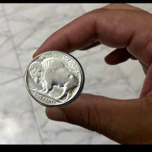 May include: A silver United States five-cent coin, featuring a detailed image of a bison on one side. The coin is held between fingers, showcasing its circular shape and the text "FIVE CENTS". The coin's design includes the words "UNITED STATES OF AMERICA".