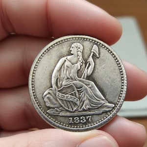 May include: A silver coin with a seated female figure holding a staff and shield, inscribed with "LIBERTY" and the date "1837". The coin has a detailed design and a circular shape, held in a hand.
