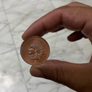 May include: A copper Indian Head penny from 1873, held between fingers. The coin features a profile of a Native American in a feathered headdress and the words "UNITED STATES OF AMERICA".