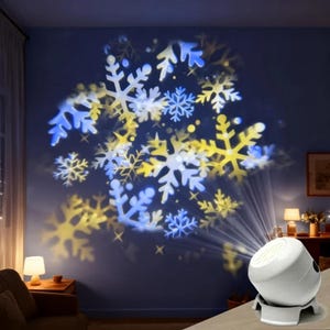 Puede incluir: Un proyector de copos de nieve blanco proyecta patrones de copos de nieve azules y amarillos sobre una pared azul oscuro. El proyector está sobre una superficie clara, con una lámpara y muebles en el fondo.