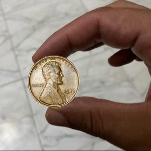 May include: A copper-colored 1943 Lincoln penny held between fingers. The coin features a profile of Abraham Lincoln and the word "LIBERTY". The year "1943" is visible on the right side of the coin.