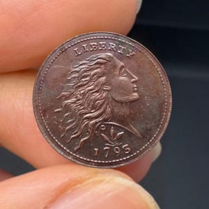 May include: A copper-colored coin featuring a profile of a woman with flowing hair, the word "LIBERTY" above, and the date "1795" below. The coin has a detailed design and a slightly worn appearance, suggesting its age.
