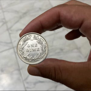 May include: A silver coin with the words "ONE DIME" in the center, surrounded by a wreath design. The coin is held between fingers, showcasing its detailed design and metallic sheen. The coin's rim has a textured edge.