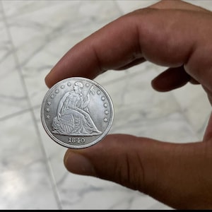 May include: A silver coin from 1840, held between fingers. The coin features a seated figure holding a staff and a shield. The coin is surrounded by stars and the year 1840 is visible at the bottom.