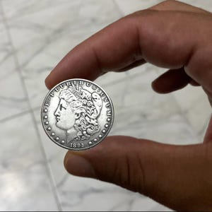 May include: A silver coin with a profile of a woman, the text "E PLURIBUS UNUM", and the year 1895. The coin is held between fingers against a marble background. The coin's details are visible, including the stars and the woman's hair.